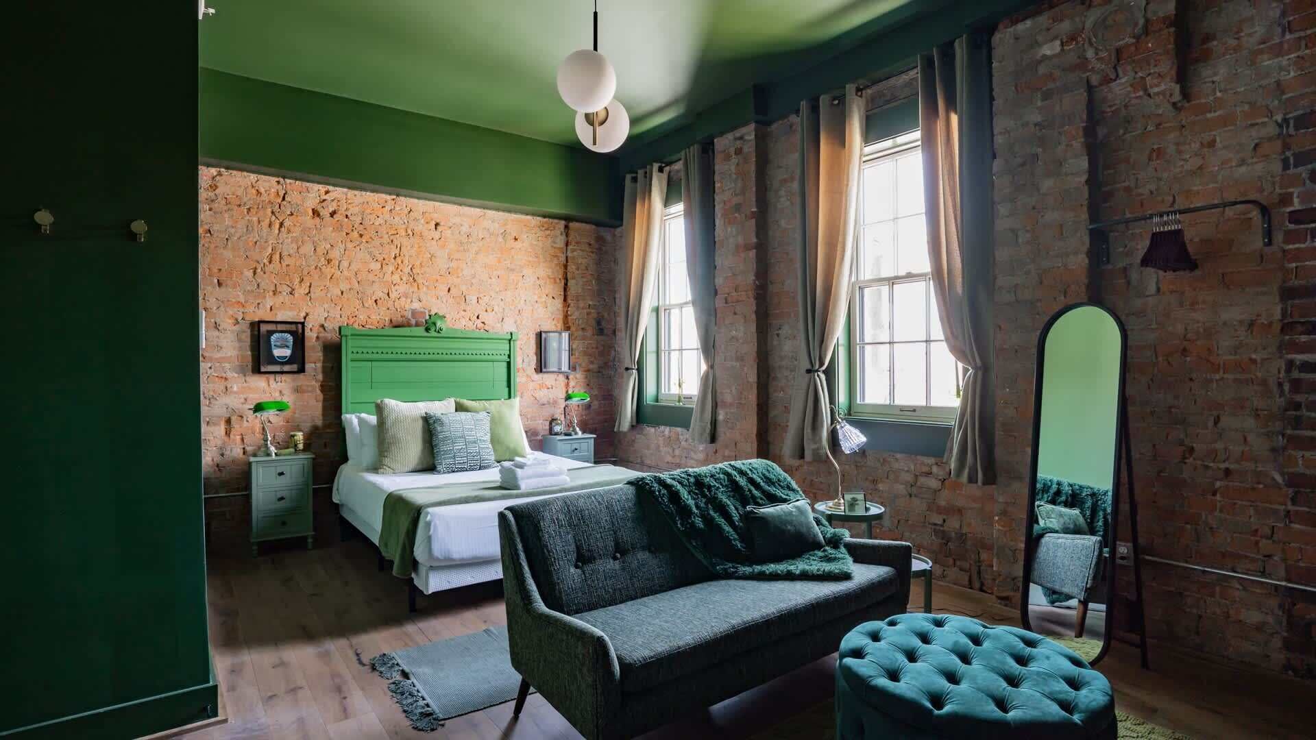 Quarto com parede verde, parede de tijolos, cama com cabeceira verde, sofá verde e um espelho.