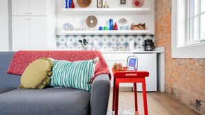 A cozy living room with a grey sofa, a red blanket, and striped pillows. A small red table with a card holder is in front of the sofa. The room has a kitchen with white cabinets and a brick wall. - Pickle Factory Hotel—1BR Beside New Distillery! (Covington)