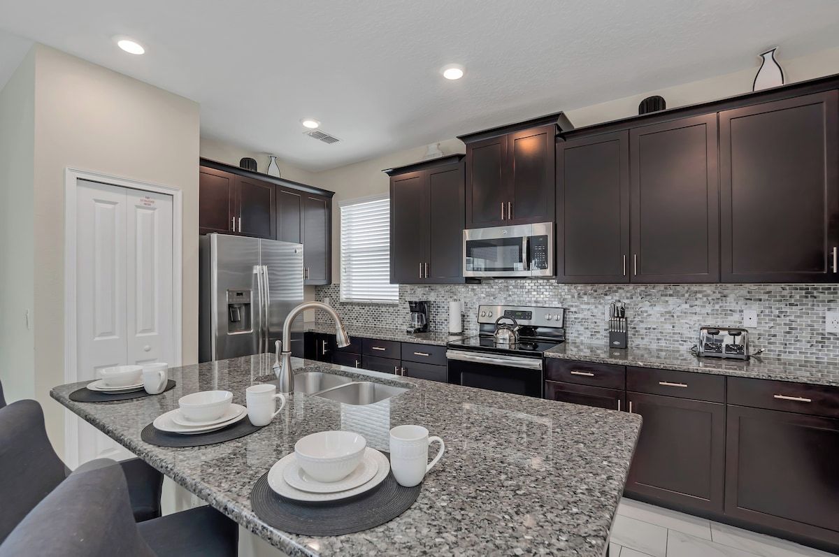 A modern kitchen with dark cabinetry, a granite countertop, stainless steel appliances, and a breakfast bar.