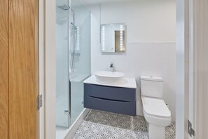 A bathroom with a glass shower enclosure, a white sink with a vessel basin, a dark blue vanity, and a white toilet. - Host & Stay | The Townhouse (Whitby)