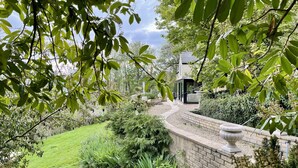 A garden with a brick wall, a pathway, and a modern house with a glass facade. - Beautiful hill side house with stunning valley view in the centre of Cotswolds (Chalford)