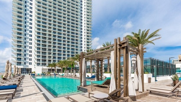 A pool area with lounge chairs and a wooden pergola, surrounded by palm trees and a tall building in the background. - Hyde Beach House Luxury Condo-resort 2 Bedroom Condo by RedAwning (Hollywood)