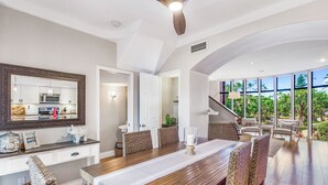 A spacious dining area with a long wooden table, wicker chairs, and a view of a lush garden through large windows. - Napili Gardens B7 3 Bedroom 3 Townhouse by RedAwning (Lahaina)