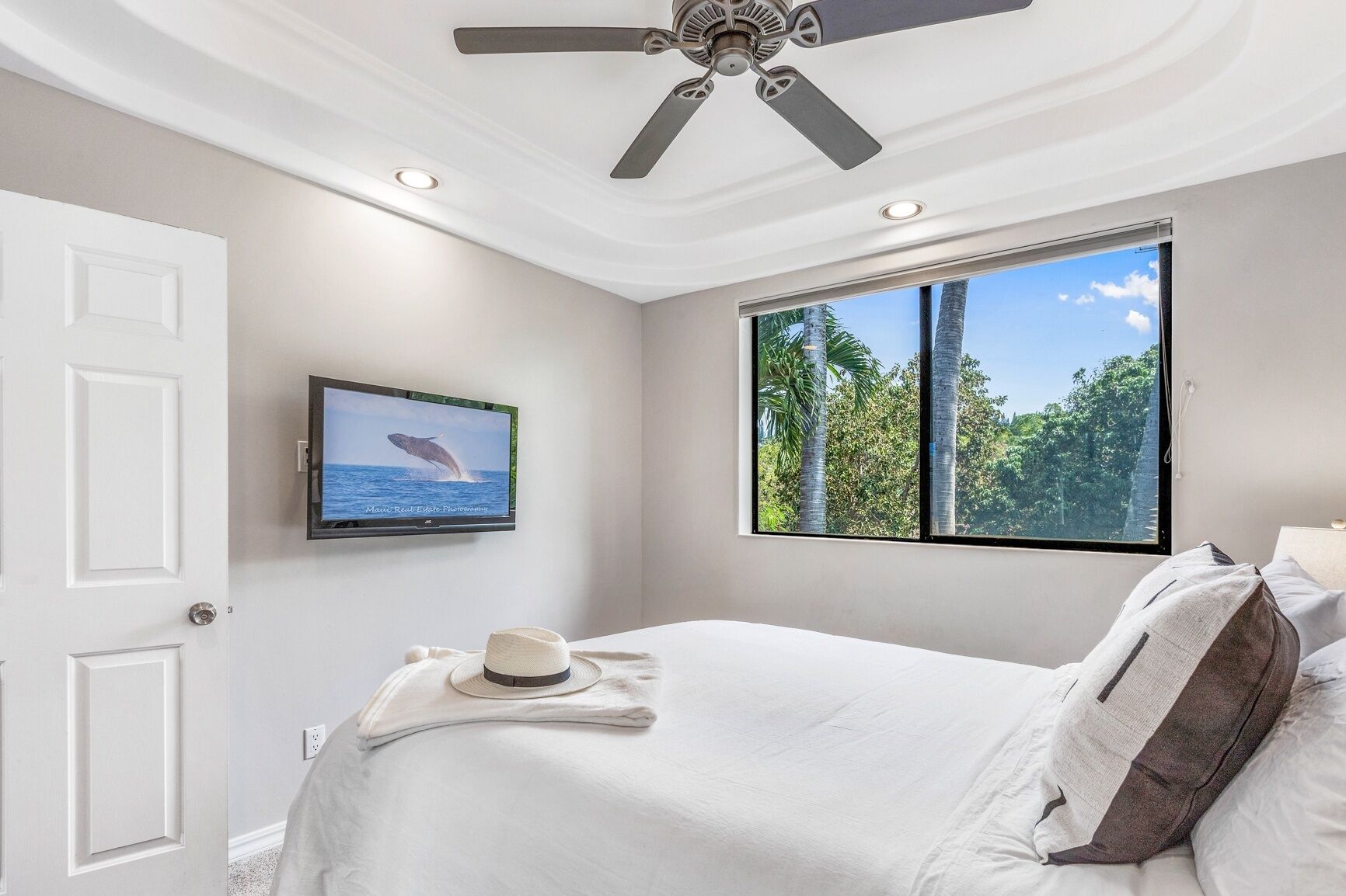 A bedroom with a bed, a ceiling fan, a TV on the wall, and a window with a view of trees.