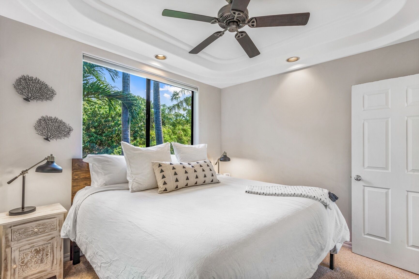A bedroom with a large bed, a ceiling fan, a window with a view of palm trees, and a white door.