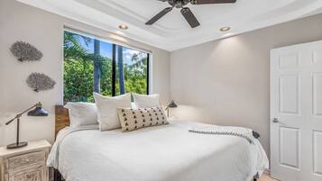 A bedroom with a large bed, a ceiling fan, a window with a view of palm trees, and a white door.