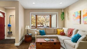 A living room with a beige sofa, colorful cushions, a wooden coffee table, a floor lamp, and a large window with a view of trees. - Whistler Village - Redwood 10 2 Bedroom Condo by RedAwning (Steamboat Springs)