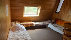 Een knus, klein kamertje met houten wanden en een schuine kap. Er staan twee bedden met kleurrijk beddengoed in, een klein raam en een houten bankje.