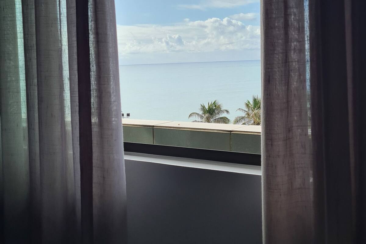 A room with a view of the sea through a window, curtains, and a table.