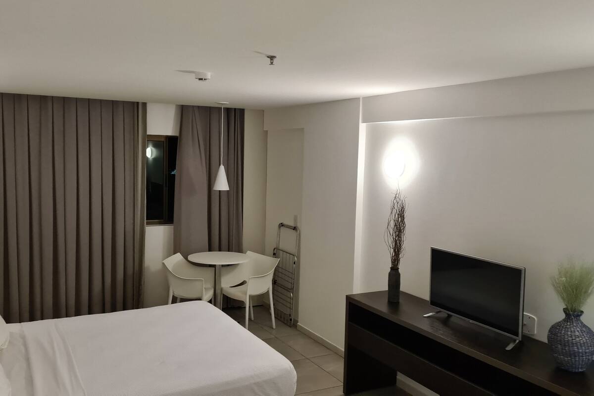 A hotel room with a bed, a desk with a TV, a chair, a small table, and a window with curtains.