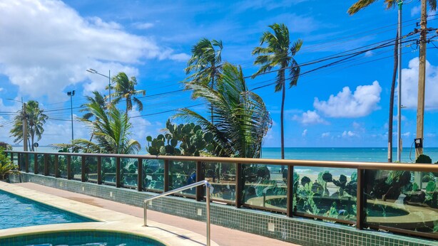 A swimming pool with a tiled edge, surrounded by palm trees and a view of the ocean. - Ritz Suítes Maceió Flat particular (Maceió)