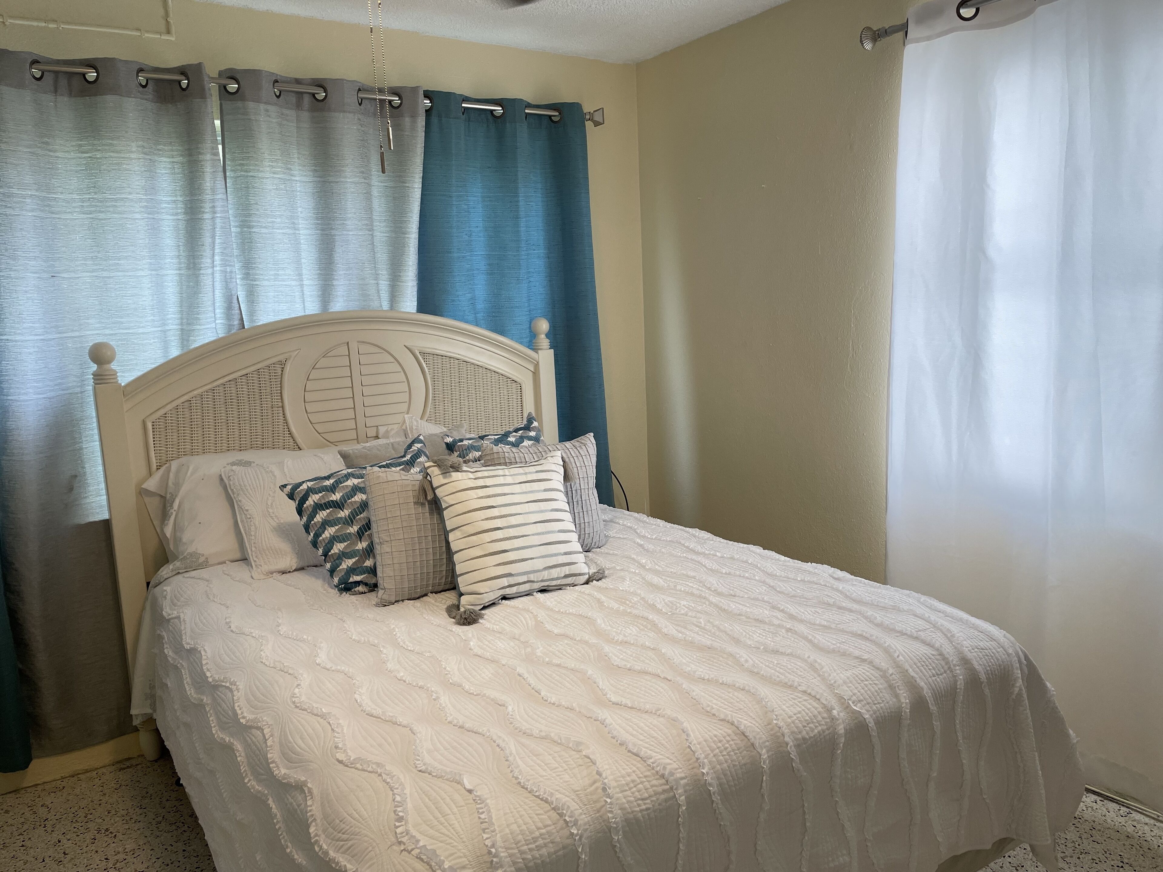 A neatly made bed with a white quilt and several pillows, a wooden headboard, and a window with a white curtain.