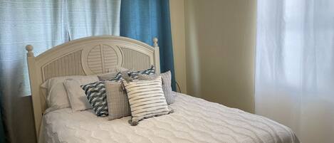 A neatly made bed with a white quilt and several pillows, a wooden headboard, and a window with a white curtain.