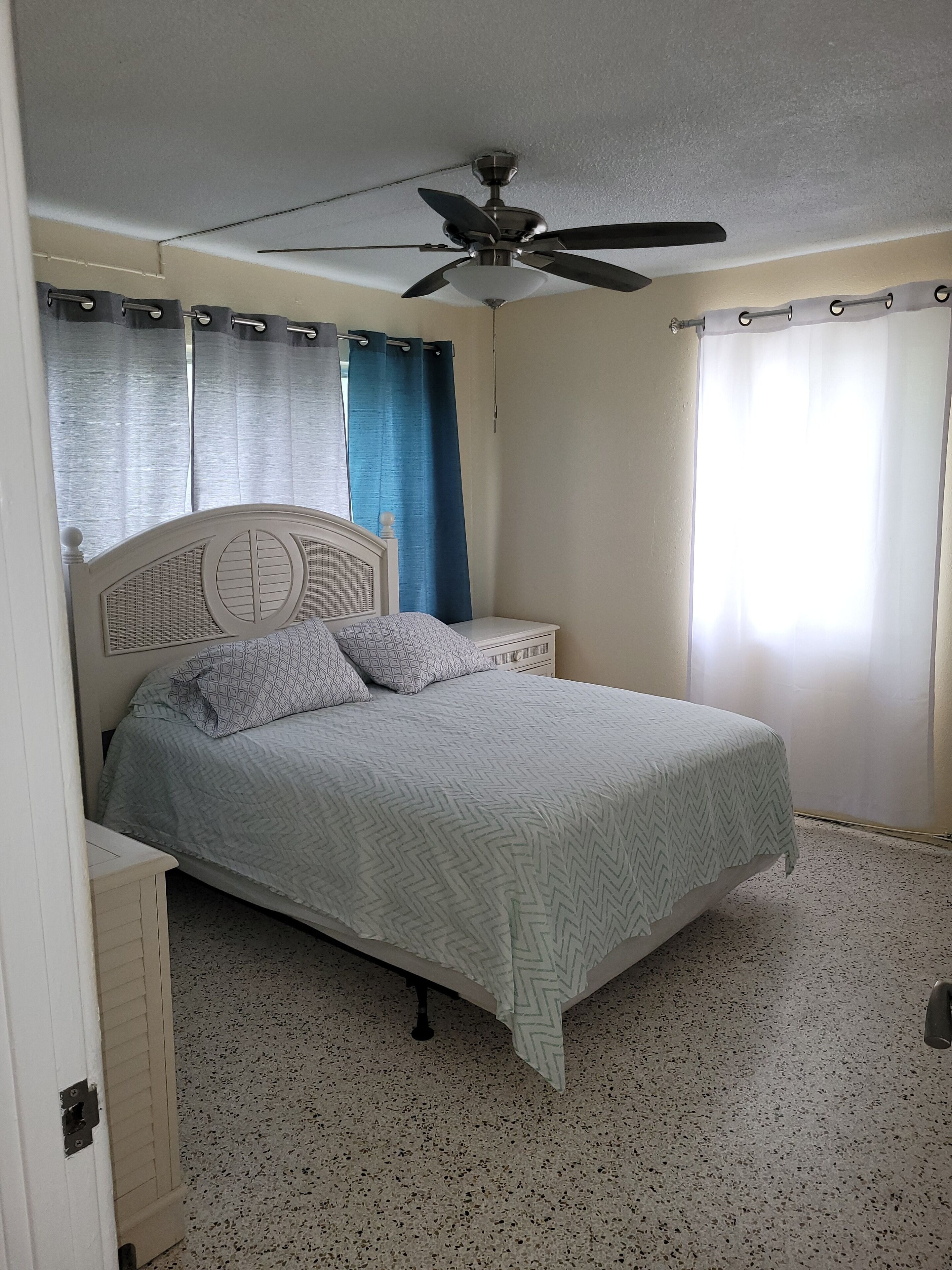 Un dormitorio con cama, ventilador de techo, mesita de noche y ventana con cortinas.