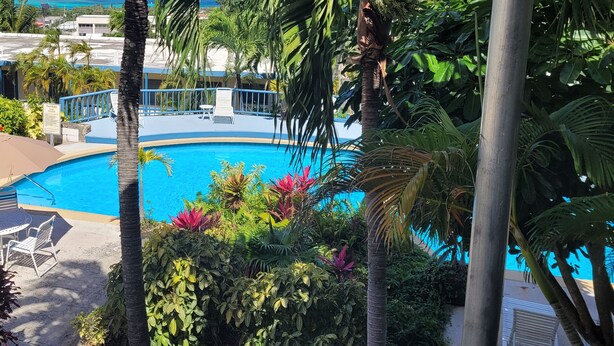A pool surrounded by tropical plants and palm trees, with a view of the ocean and buildings in the background. - The Island Life Condo@ Long Reef with Separate Bedroom + True Ocean View (Christiansted)