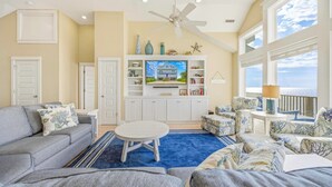 A spacious living room with a large TV, comfortable seating, and a view of the ocean. - Soundfront w/ Pool, Hot Tub, Elevator, Game and Media Rooms, Windsports Access (Avon)