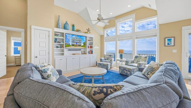 A spacious living room with a large gray sofa, a round coffee table, and a ceiling fan. There is a view of the ocean through large windows. - Soundfront w/ Pool, Hot Tub, Elevator, Game and Media Rooms, Windsports Access (Avon)
