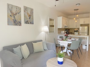 Una sala moderna con un sofá gris, una mesa de comedor preparada para cuatro personas y una cocina con armarios blancos y un refrigerador.