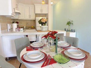 Comedor con una mesa redonda preparada para cuatro personas, armarios blancos, una cocina al fondo y una planta en maceta sobre la encimera.