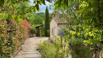 Un chemin de gravier mène à un bâtiment doté d’un toit en tuiles, entouré d’une végétation luxuriante et de diverses plantes.