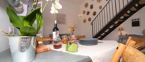 Ein Essbereich mit Tisch für zwei Personen, einem Topf mit Orchideen, einer Weinflasche und einer Obstschale. Im Hintergrund ist eine Treppe zu sehen.