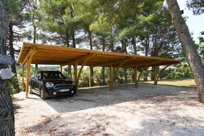 A covered parking area with a car parked under a wooden structure, surrounded by trees.