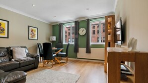 A living room with a sofa, a dining table, chairs, a clock, and a view of a building through a window. - 5 St Georges House · Lovely 1-Bed Apartment Right in the Heart of York - Pass the Keys (York)
