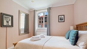 A neatly made bed with towels, a window with curtains, and framed pictures on the walls. - 5 St Georges House · Lovely 1-Bed Apartment Right in the Heart of York - Pass the Keys (York)