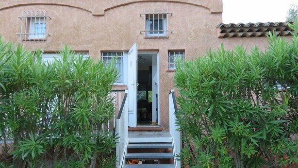 Un bâtiment à l’extérieur de couleur beige, avec un enduit texturé, un escalier menant à une porte ouverte et de la verdure tout autour.