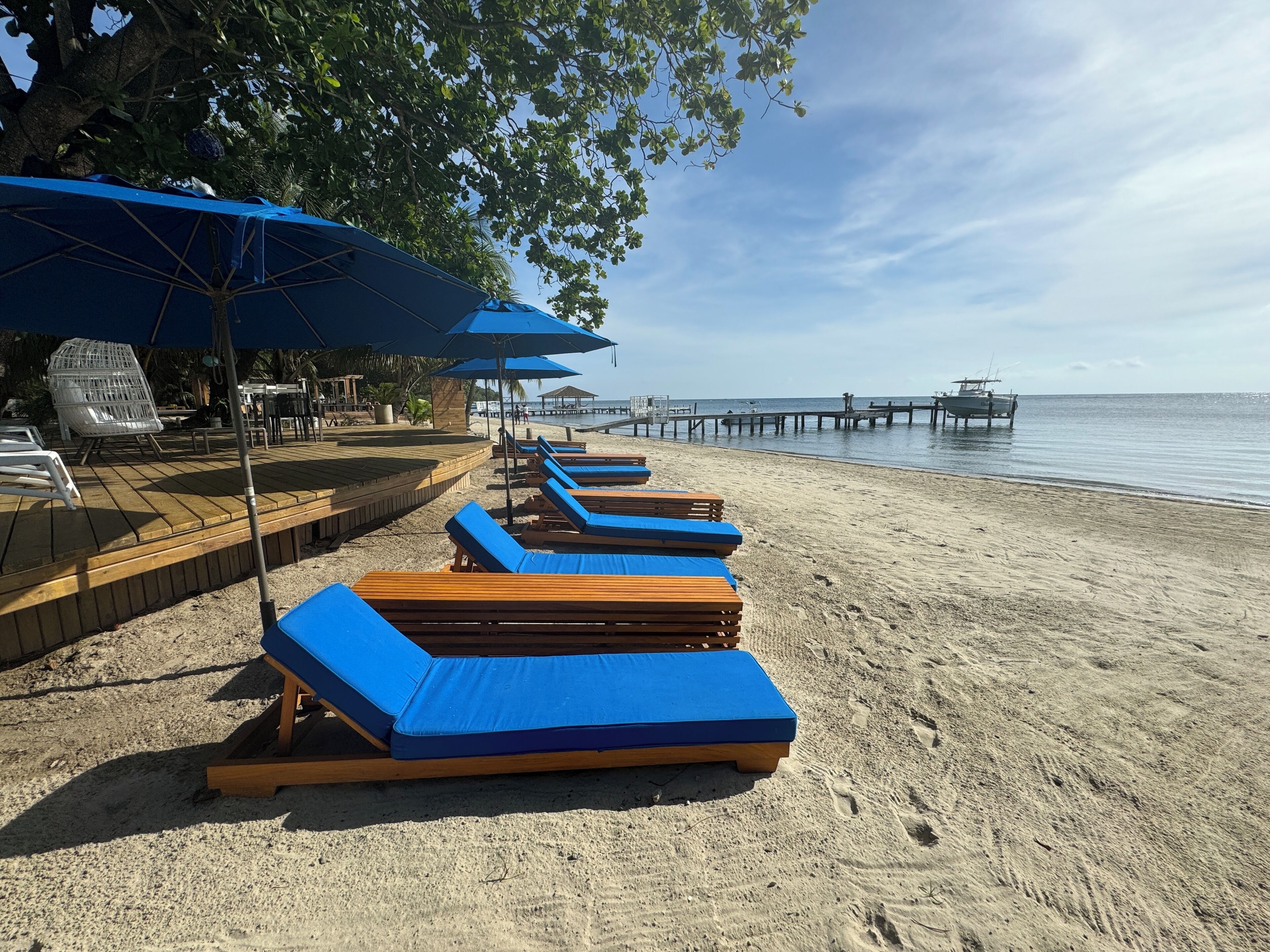 A beachside lounge area with wooden loungers and blue cushions, a wooden deck, and a pier extending into the water.