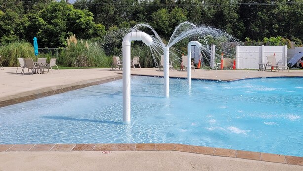 A swimming pool with two water jets, surrounded by lounge chairs and umbrellas, with a fence and greenery in the background. - Breezy Beach Retreat (Gulfport)