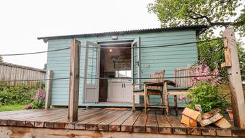 A rustic cabin with a wooden deck, a dining area with chairs, and a small kitchen visible through an open door.