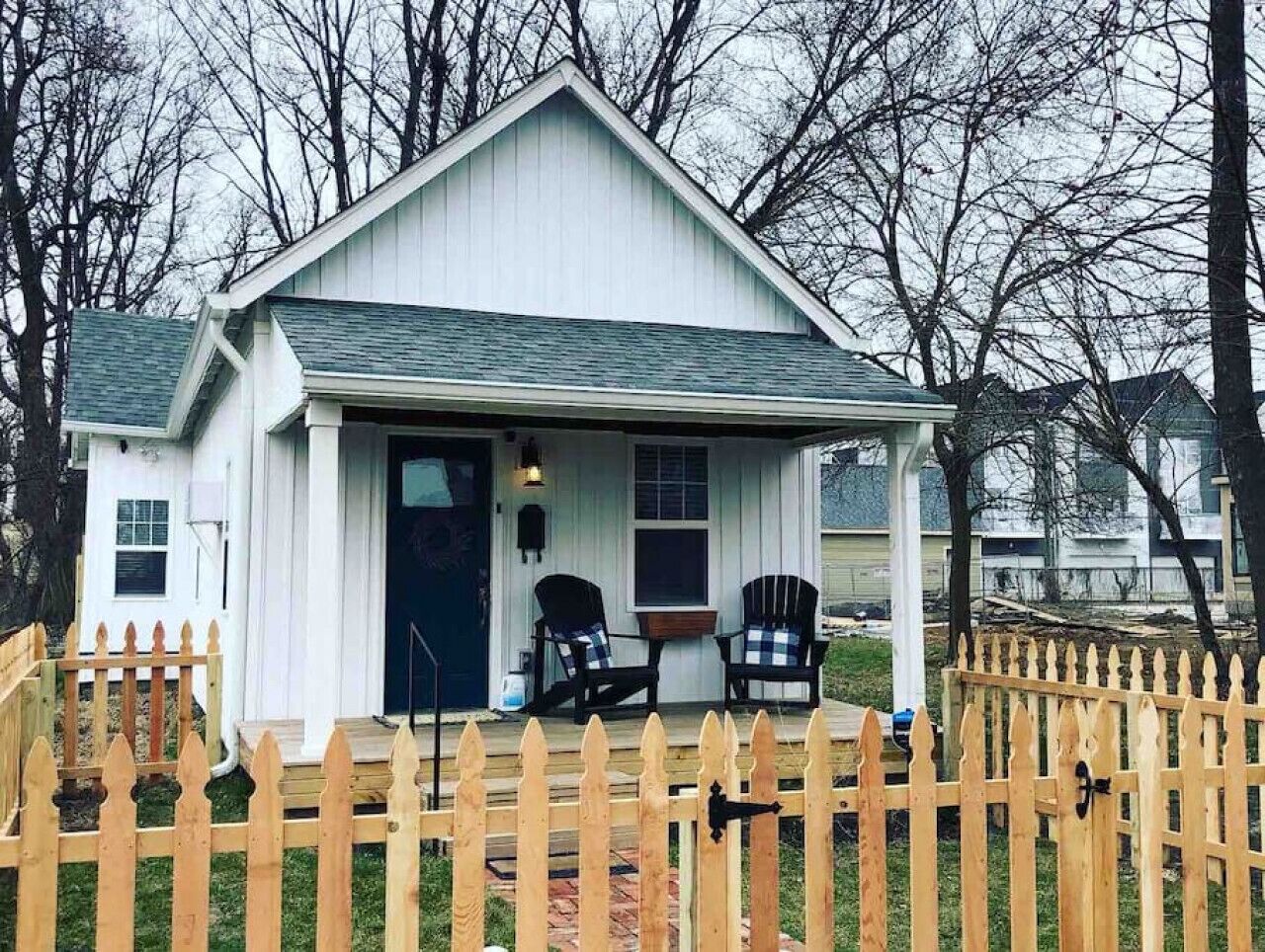 A small white house with a porch, two Adirondack chairs, and a wooden picket fence.