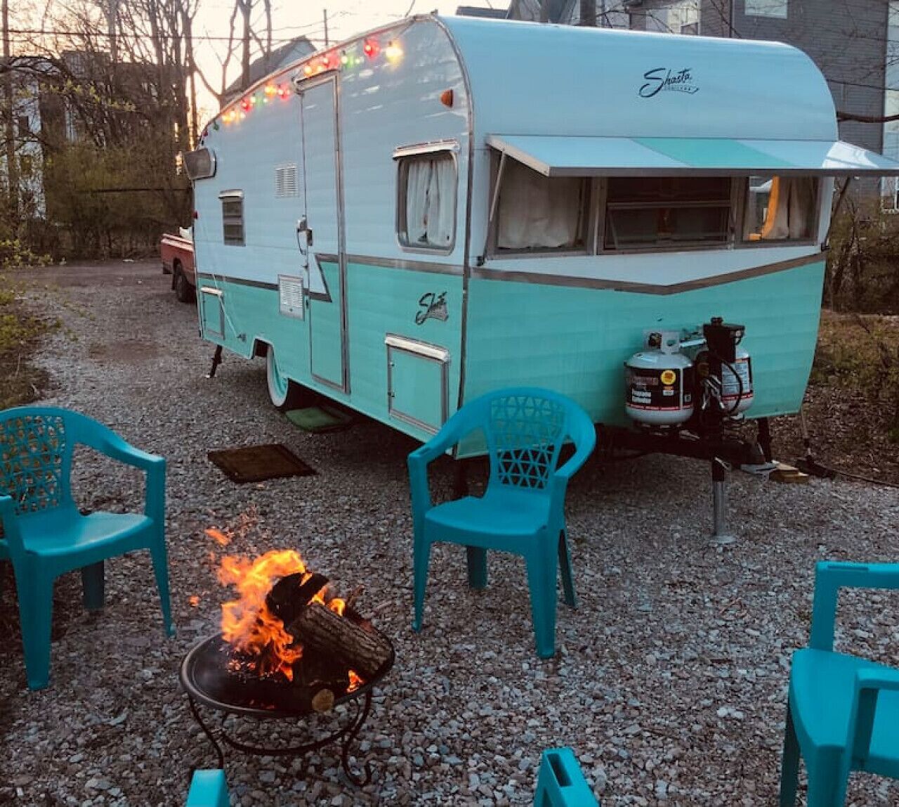 A vintage Shasta trailer with a fire pit and turquoise chairs in a gravel area.