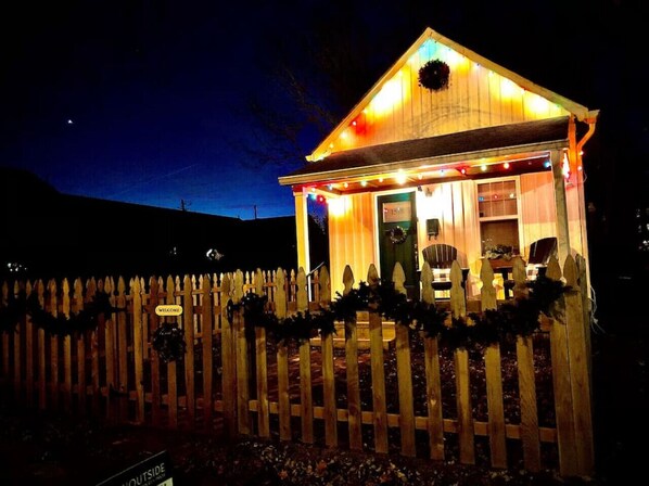 A small, illuminated wooden structure with a welcoming sign, a porch, and a picket fence.