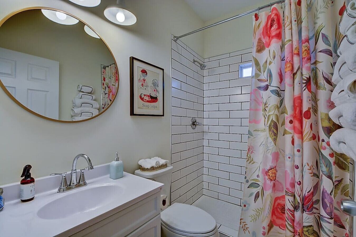 Ett badrum med ett vitt handfat, en spegel, en dusch med ett duschdraperi med blommotiv och en toalett.