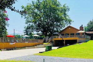 A building with a wooden exterior, a covered outdoor area with umbrellas, and a large tree in front.