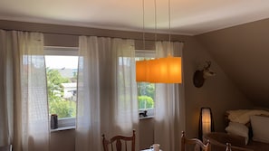 A room with a dining table, chairs, a hanging orange light fixture, a mounted deer head, and a window with sheer curtains.