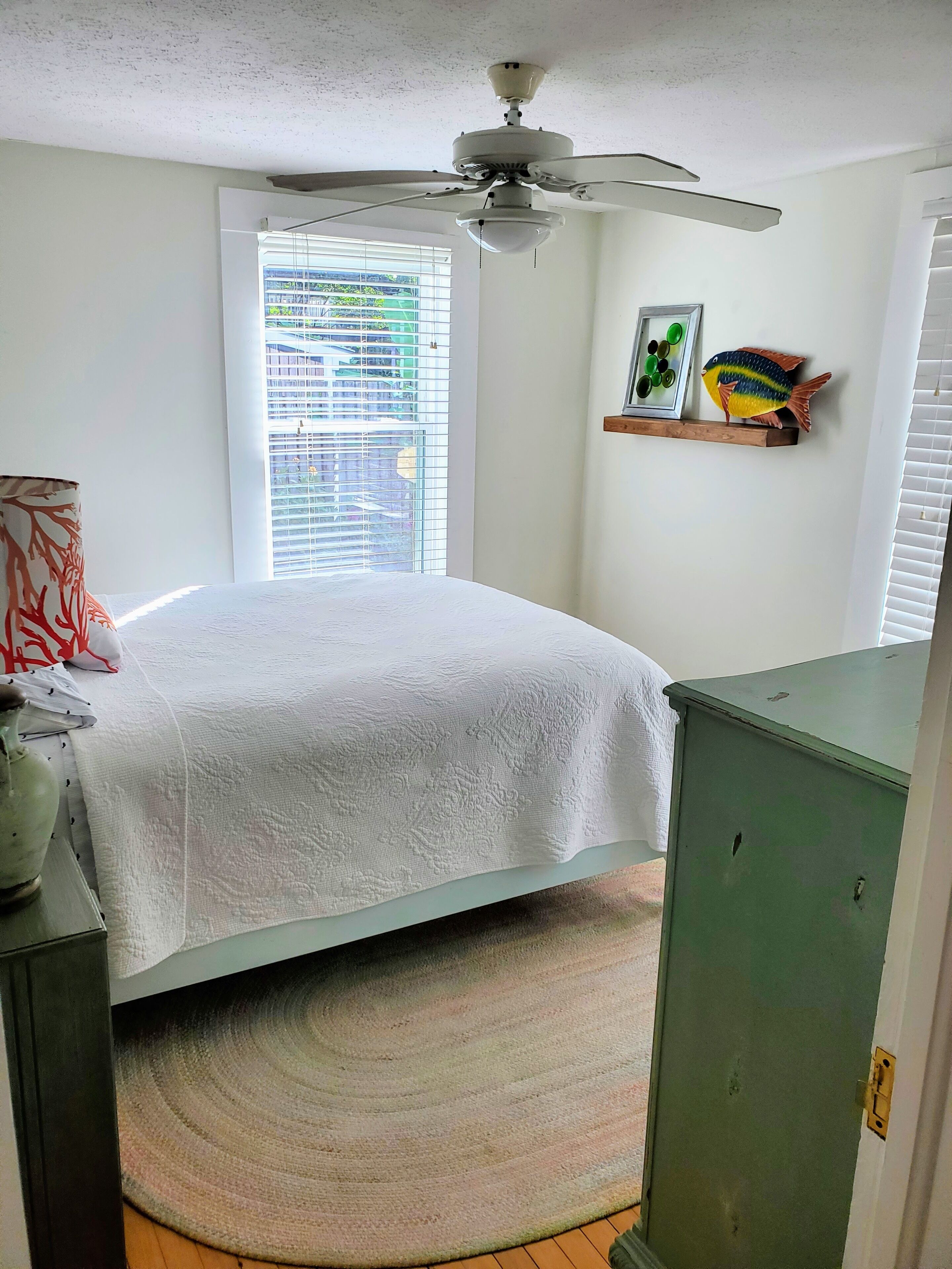 A bedroom with a bed, a ceiling fan, a window with blinds, a green cabinet, and a shelf with decorative items.