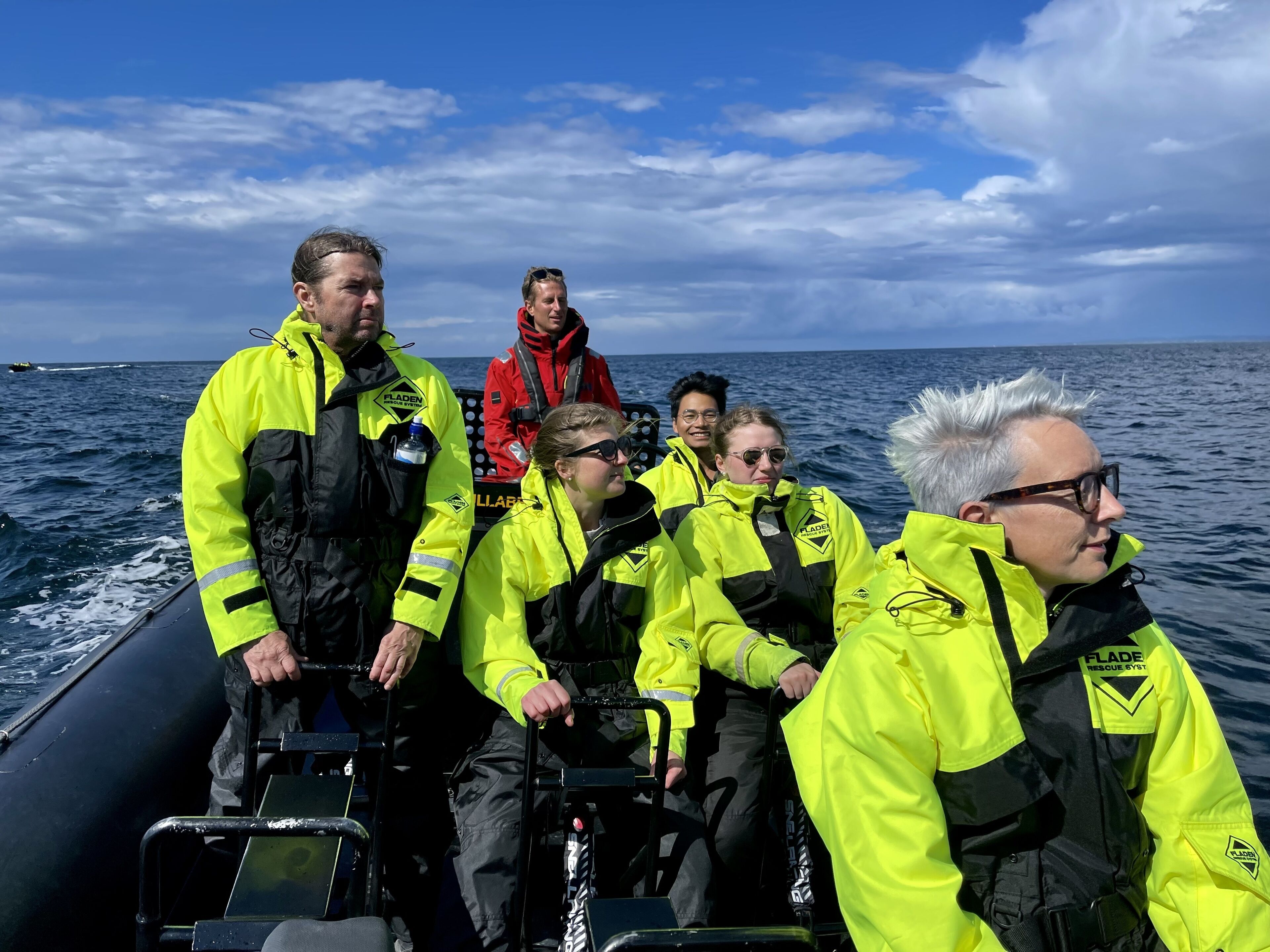 Un grupo de personas en un bote, vistiendo chaquetas amarillo brillante, algunas con pantalones negros y una con chaqueta roja.