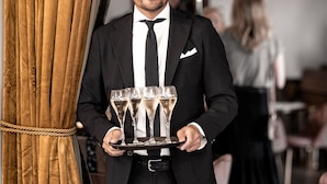 A man in a black suit holding a tray with glasses, standing in a room with a large curtain and a chandelier. - Villa Brunnby (Nyhamnslage)