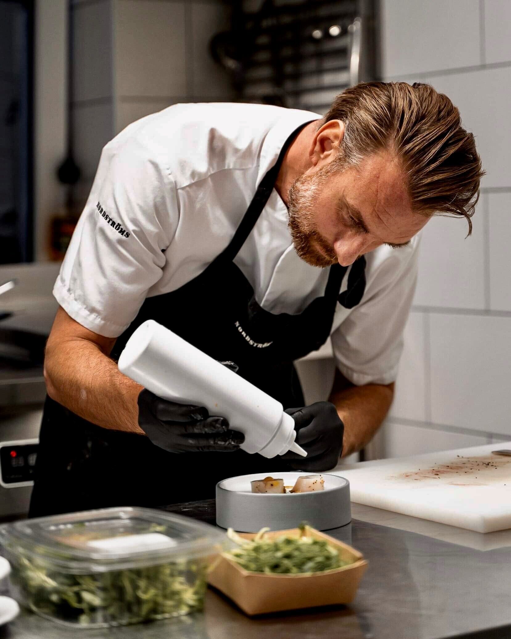 Un chef con camisa blanca y delantal negro preparando comida en una cocina.