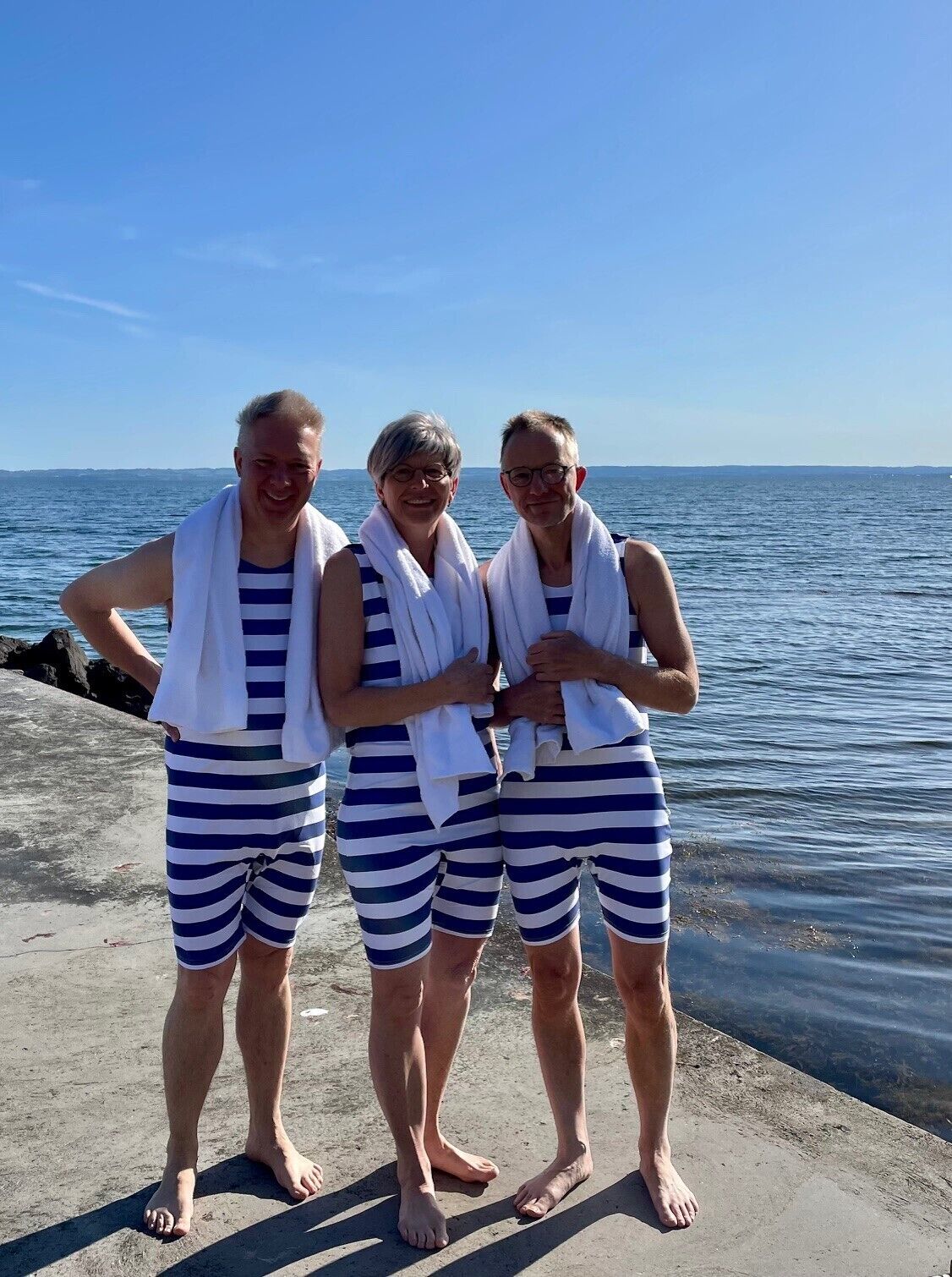 Three people standing on a concrete platform by the sea, wearing striped swimwear and white towels.