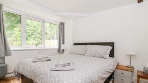 A bedroom with a bed, a nightstand, a lamp, a window with curtains, and a view of greenery outside. - Claire Cottage (Selby)