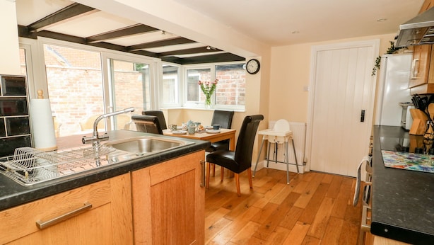 A modern kitchen with wooden cabinets, a black countertop, and a dining area with a high chair. - Claire Cottage (Selby)