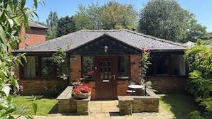 A cottage with a brick exterior, a wooden door, and a small garden with a stone bench and flower pots. - Claire Cottage (Selby)