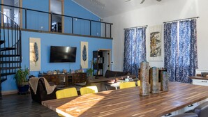 Ein geräumiger Wohnbereich mit einem Holzesstisch, gelben Stühlen, einem schwarzen Sofa und einem an der Wand montierten Fernseher.