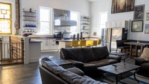 Eine moderne Küche mit weißen Schränken, einer Edelstahl-Dunstabzugshaube und einem eingebauten Backofen. Es gibt einen Essbereich mit einem gelben Stuhl und einem schwarzen Ledersofa. Der Raum verfügt über ein großes Fenster und eine städtische Aussicht.