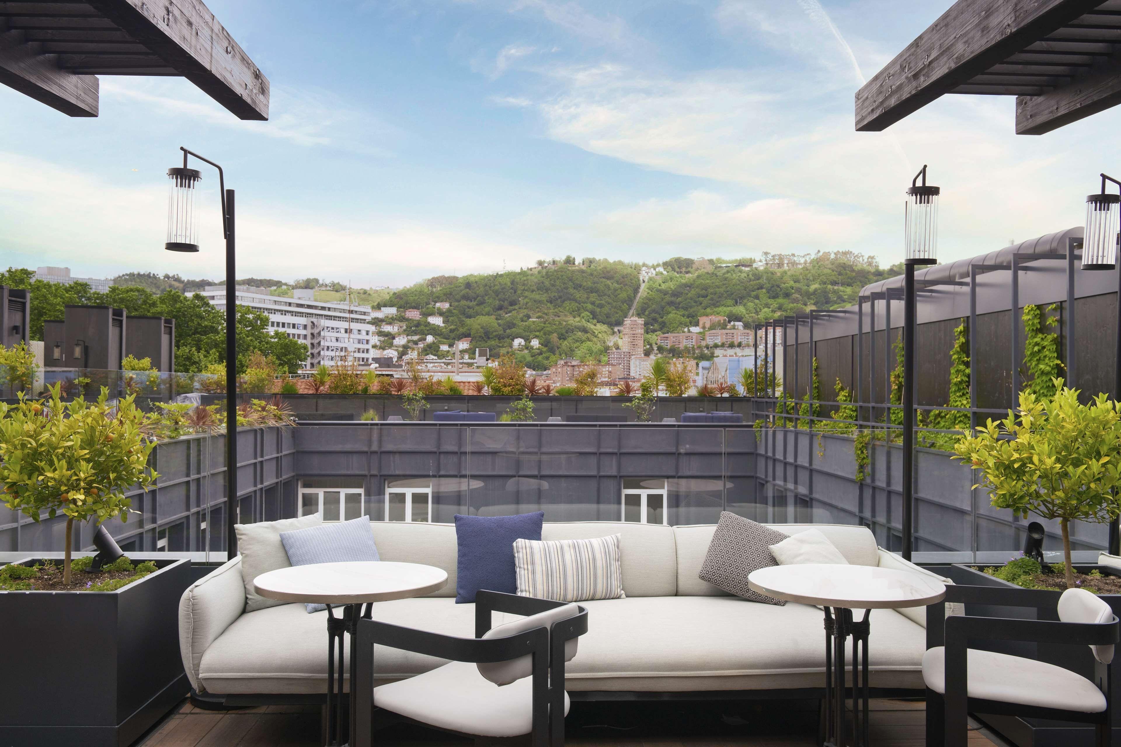 An outdoor seating area with a sofa and tables, surrounded by greenery and overlooking a cityscape.