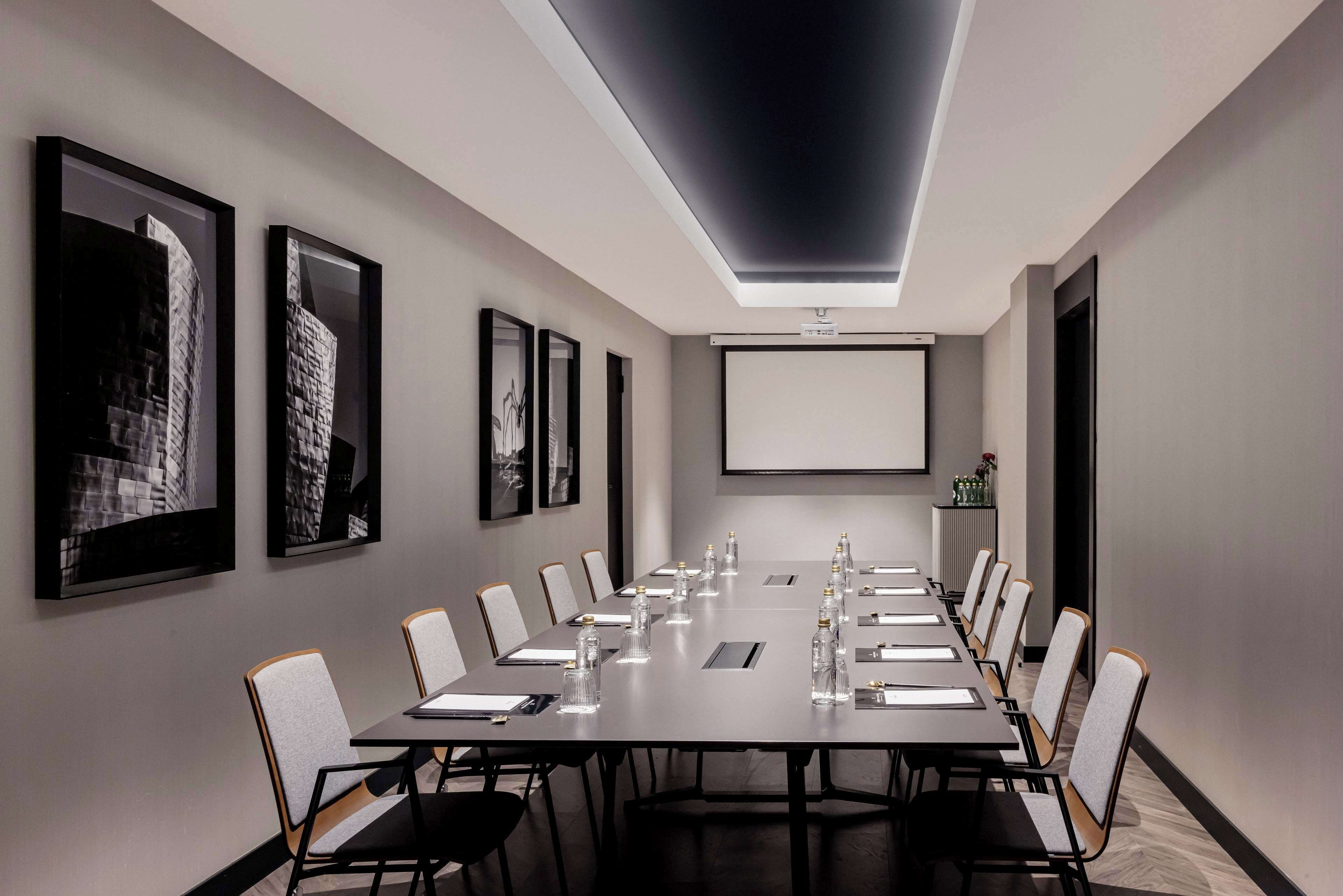 A conference room with a long table, chairs, a projector screen, and framed pictures on the walls.
